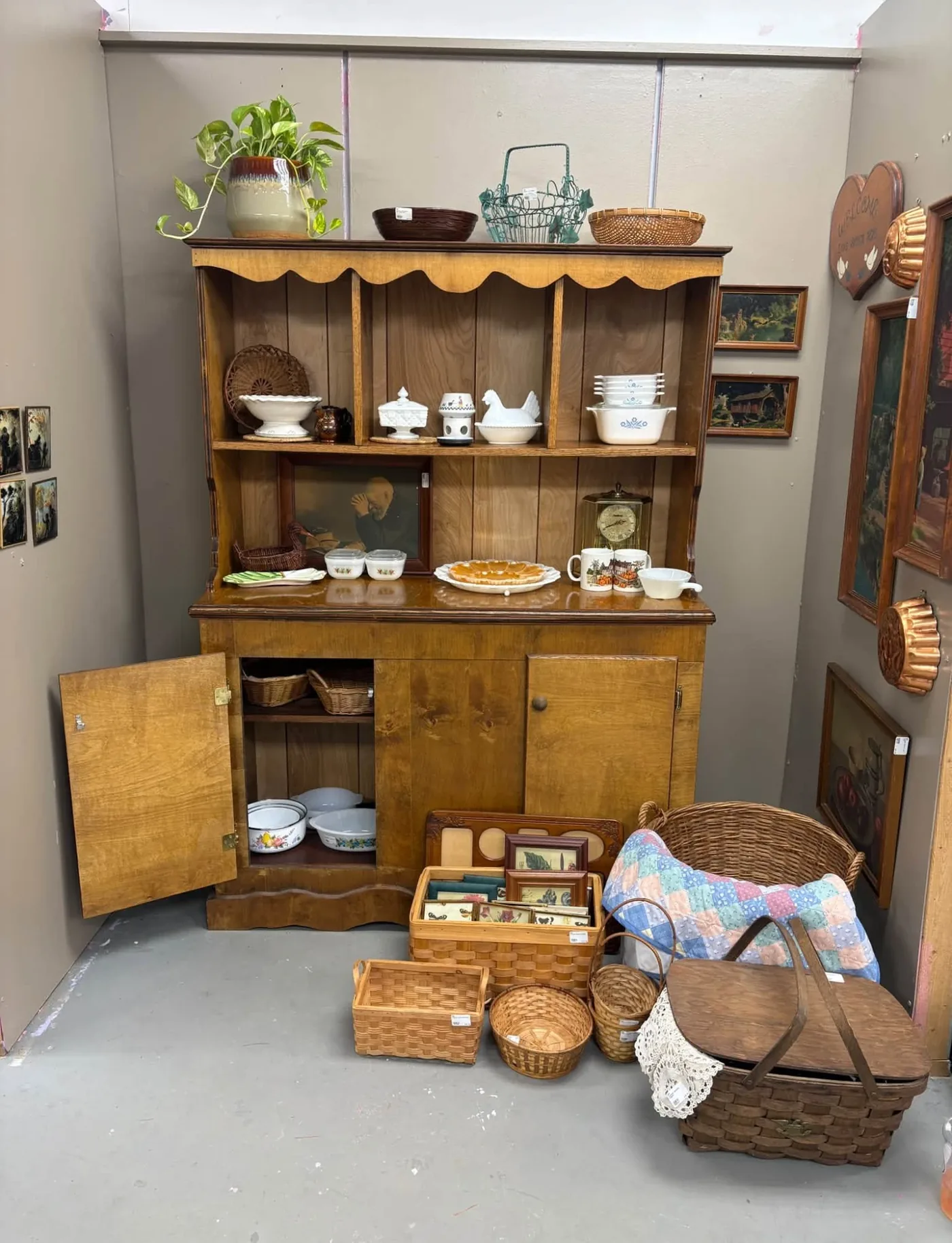 The Meemaw & Peepaw Co. booth: a honey colored wooden hutch filled with milk glass, Corning Ware and baskets, framed pictures on the walls and a patchwork quilt in a basket